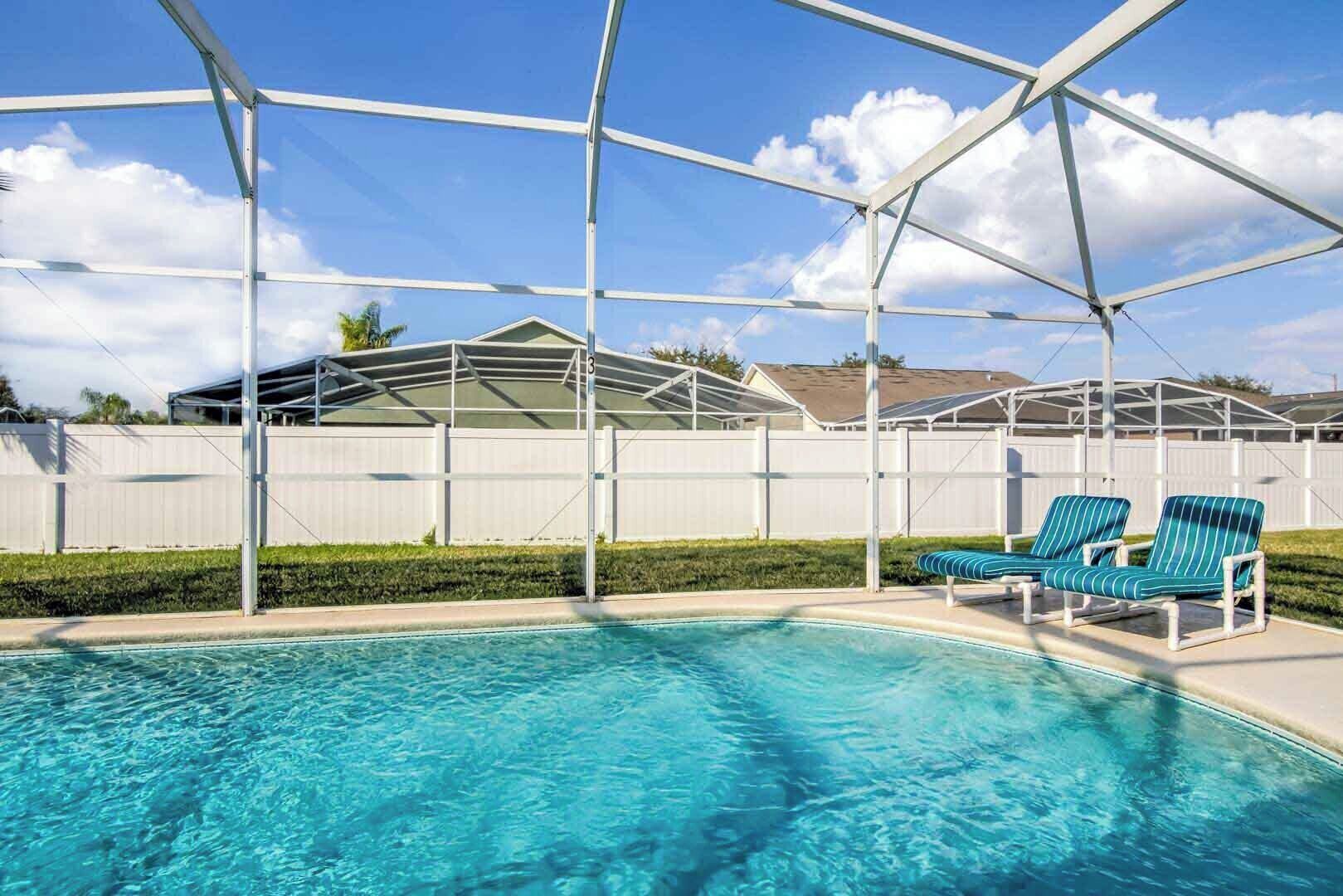 A swimming pool with a covered area, two lounge chairs, and a white fence in the background.