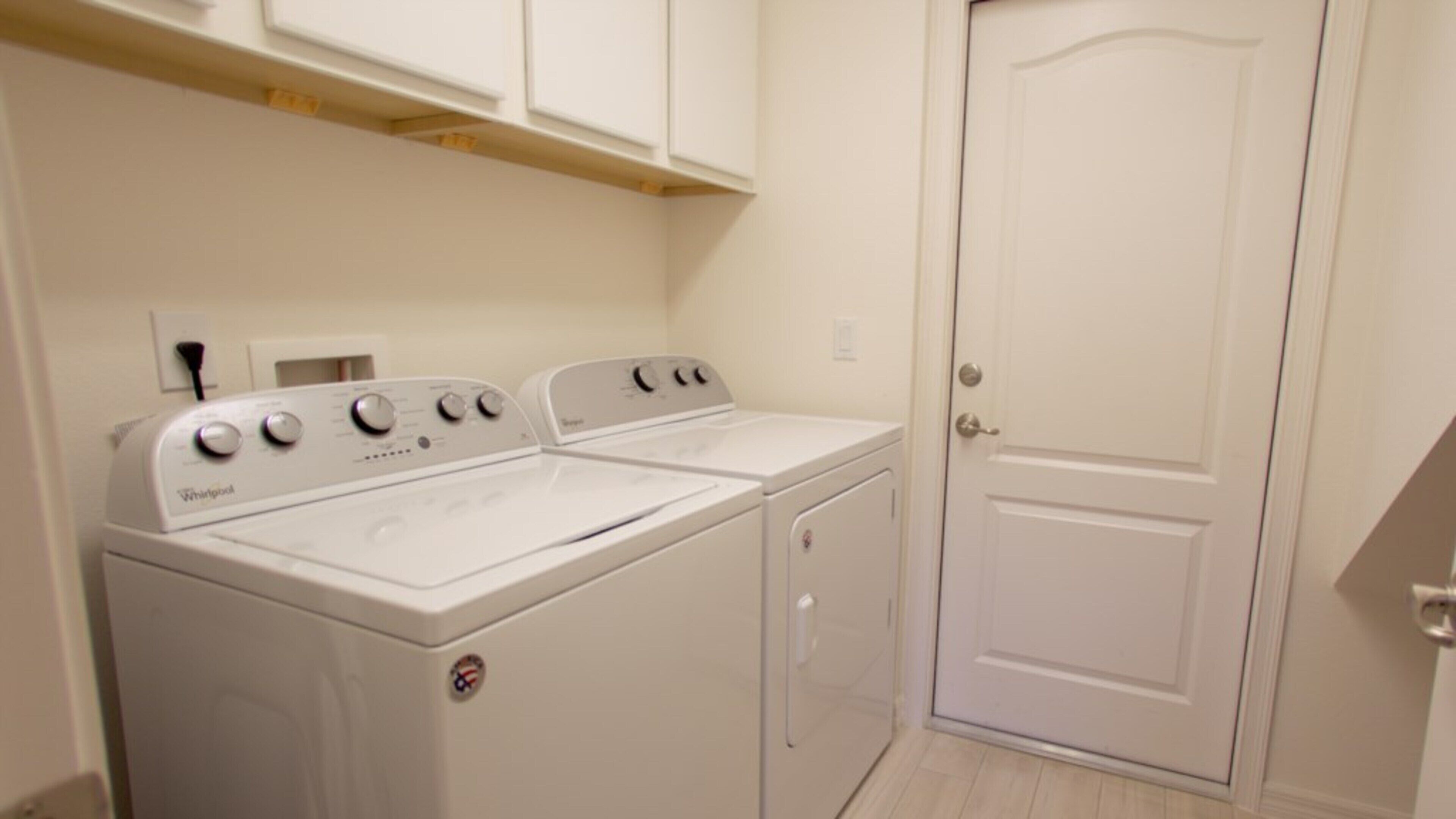 Une buanderie équipée d’un lave-linge et d’un sèche-linge Whirlpool, d’une porte blanche et d’une étagère en bois au-dessus du lave-linge.