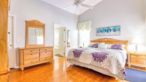 Une chambre avec une commode en bois, un lit recouvert d’une courtepointe à motifs floraux, un ventilateur de plafond et un tableau accroché au mur.