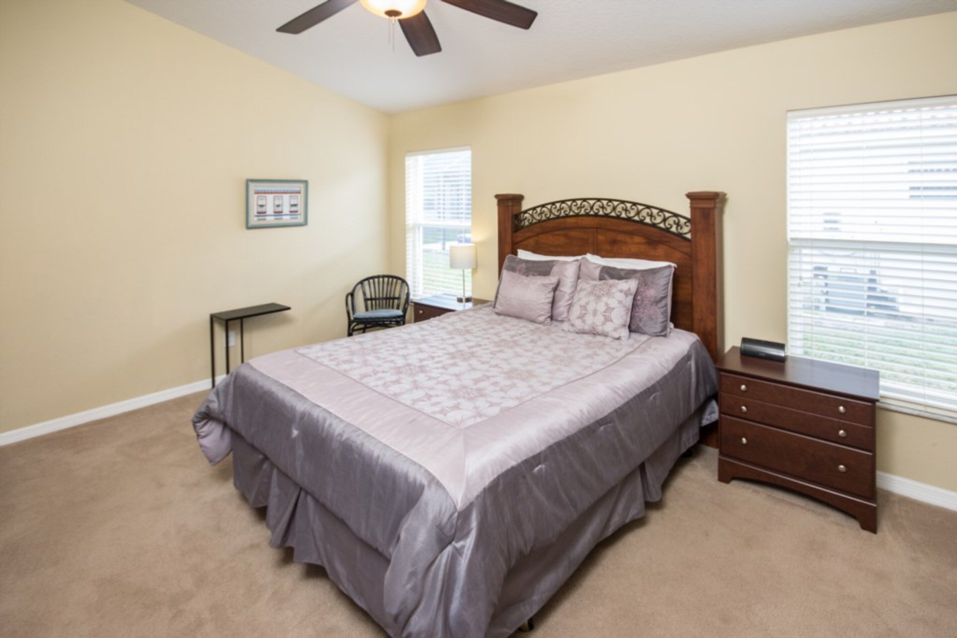 A bedroom with a large bed, a nightstand, a chair, a ceiling fan, and a window with blinds.