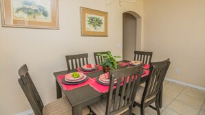 Un coin repas avec une table dressée pour quatre personnes, entourée de chaises en bois. Deux œuvres d’art encadrées ornent le mur, un lustre est suspendu au-dessus de la table et une plante en pot est posée sur la table.