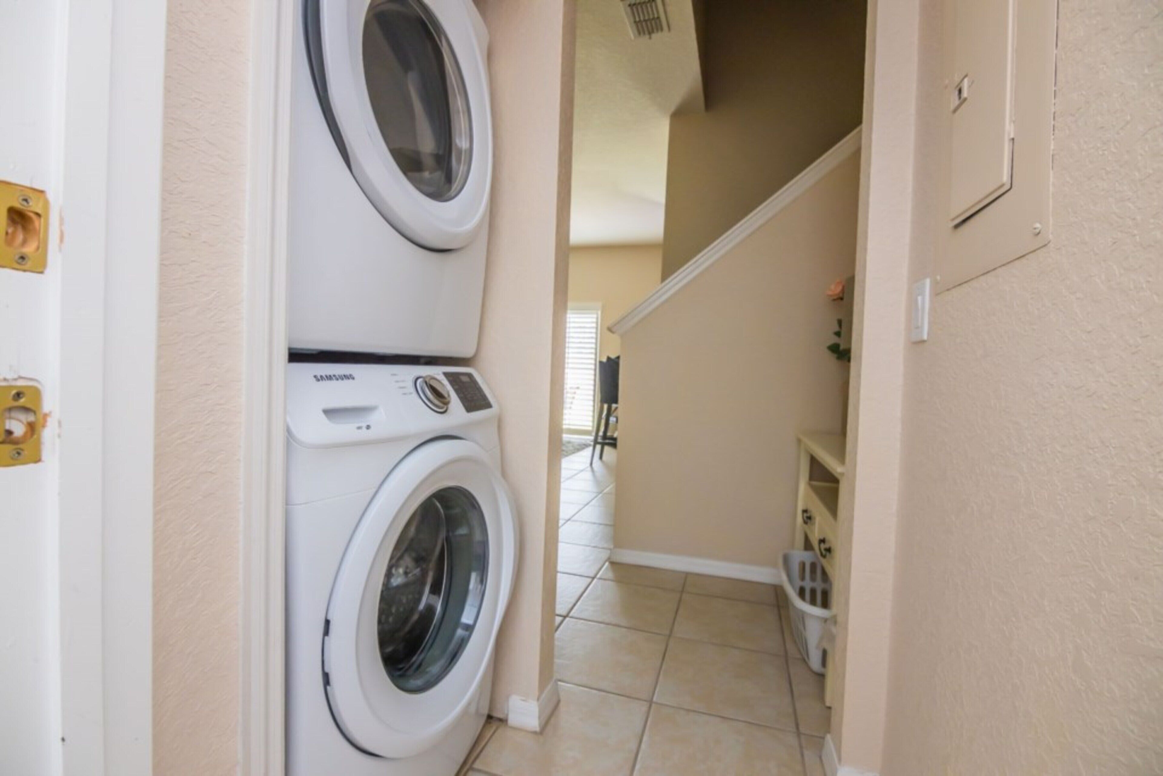 Une buanderie équipée d’un lave-linge et d’un sèche-linge Samsung, d’une petite poubelle et d’un escalier menant à une autre pièce.