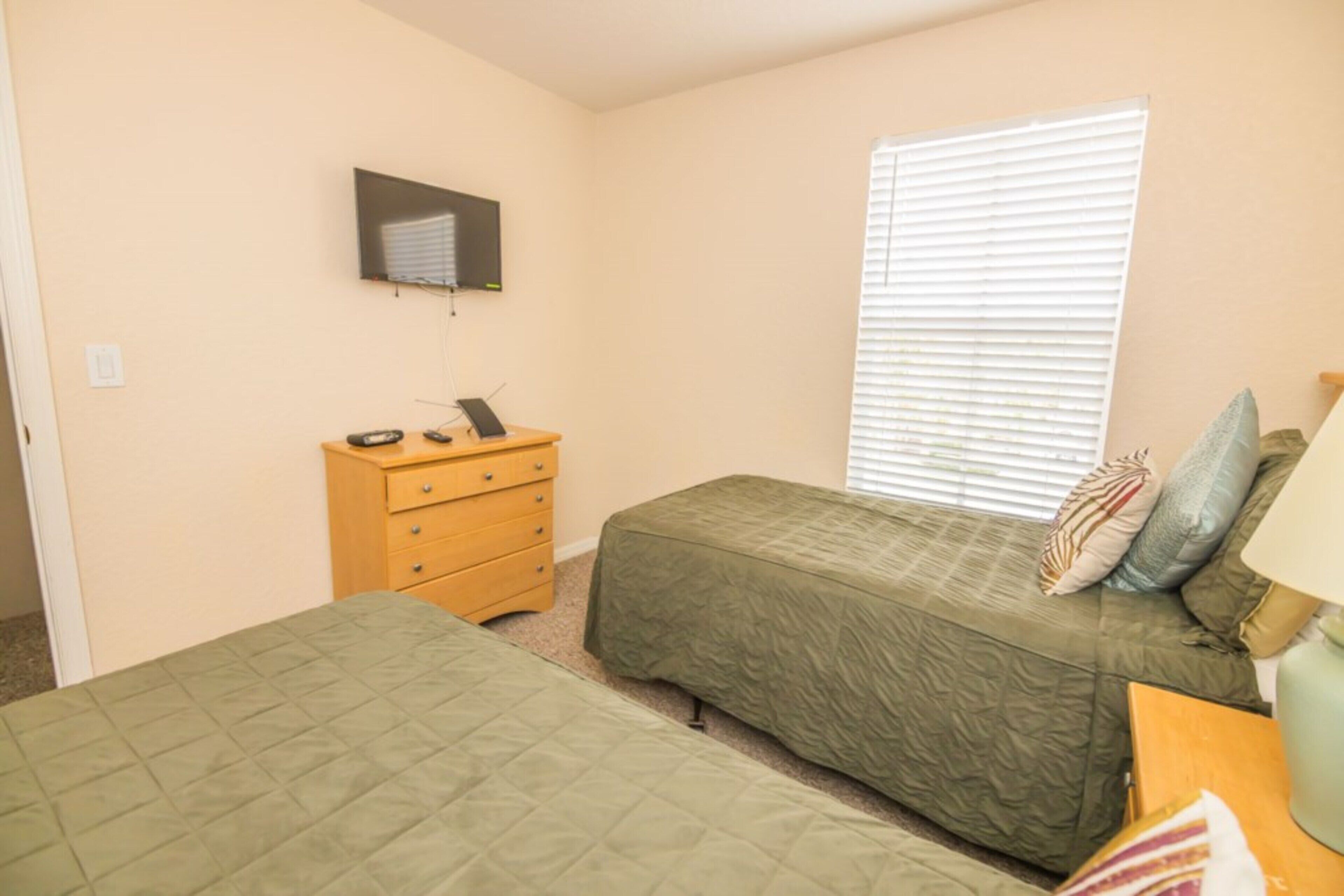 A bedroom with two beds, a wooden dresser, a television mounted on the wall, and a window with blinds.