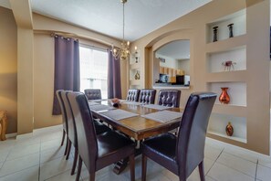 A dining room with a large wooden table, leather chairs, and a chandelier. There are shelves with decorative items on the wall.
