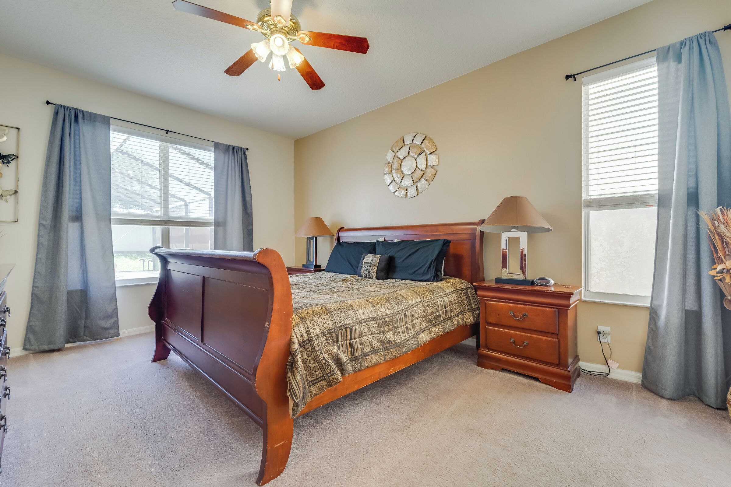 A bedroom with a wooden bed, a nightstand, a ceiling fan, a wall clock, and a window with curtains.