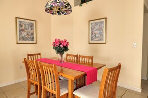 A dining room with a wooden table, six chairs, a vase of pink flowers, and two framed pictures on the walls. - Arler House - Amazing 4 Bed 3 Bath - Private Pool (Clermont)