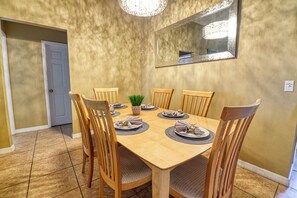 A dining room with a wooden table set for six, surrounded by wooden chairs. A small potted plant is on the table, and a mirror hangs on the wall above.