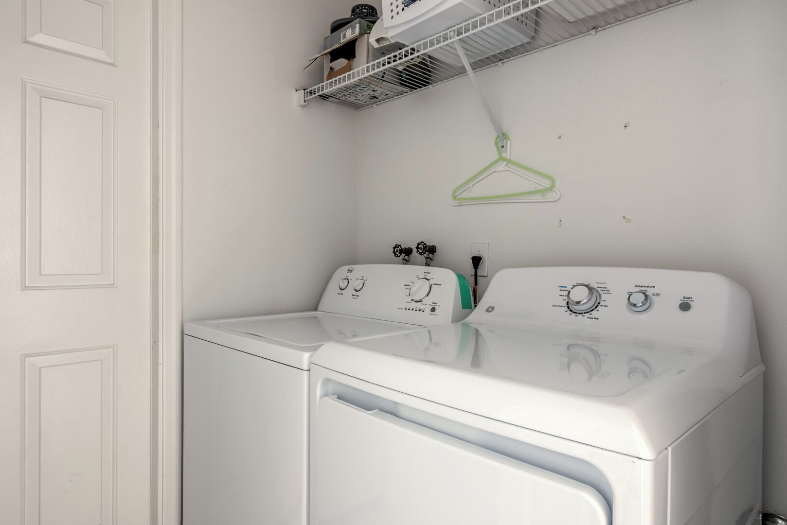 A white front-loading washer and dryer in a laundry room with a white door and a shelf above holding a green hanger.
