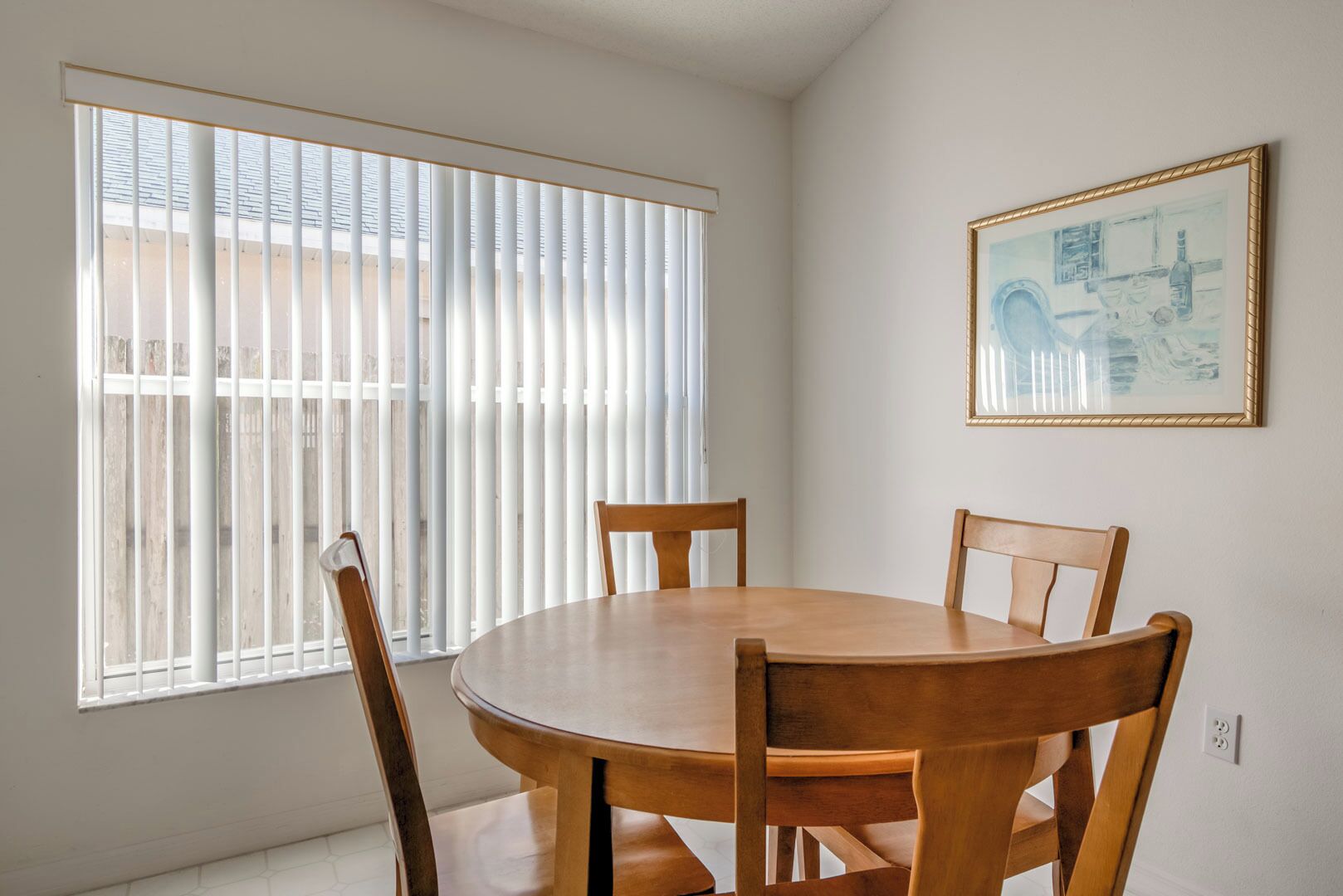 Comedor con mesa redonda de madera y cuatro sillas, ventanas amplias con persianas y un cuadro enmarcado en la pared.