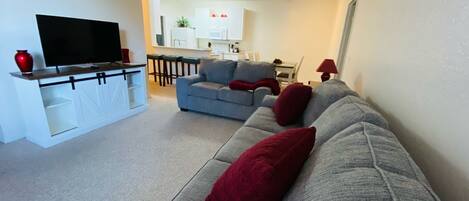 A living room with a grey sofa, red cushions, a TV on a stand, and a kitchen in the background.