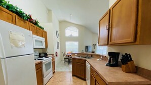Keittiössä on puukehikoiset kaapit, valkoinen jääkaappi, liesi, mikroaaltouuni ja kahvinkeitin. Taustalla näkyy ruokailutila, jossa on pöytä ja tuolit.