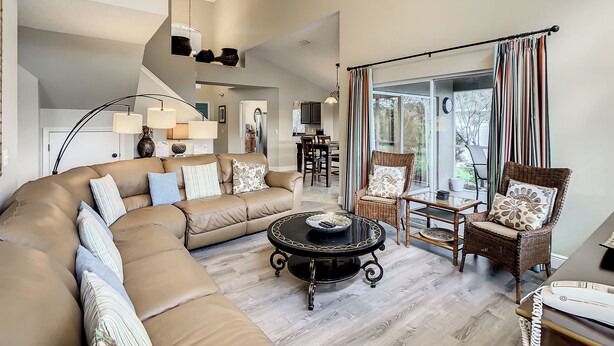 A spacious living area with a curved sofa, a round coffee table, and a wicker chair. A staircase leads to an upper level, and a dining area is visible in the background.
