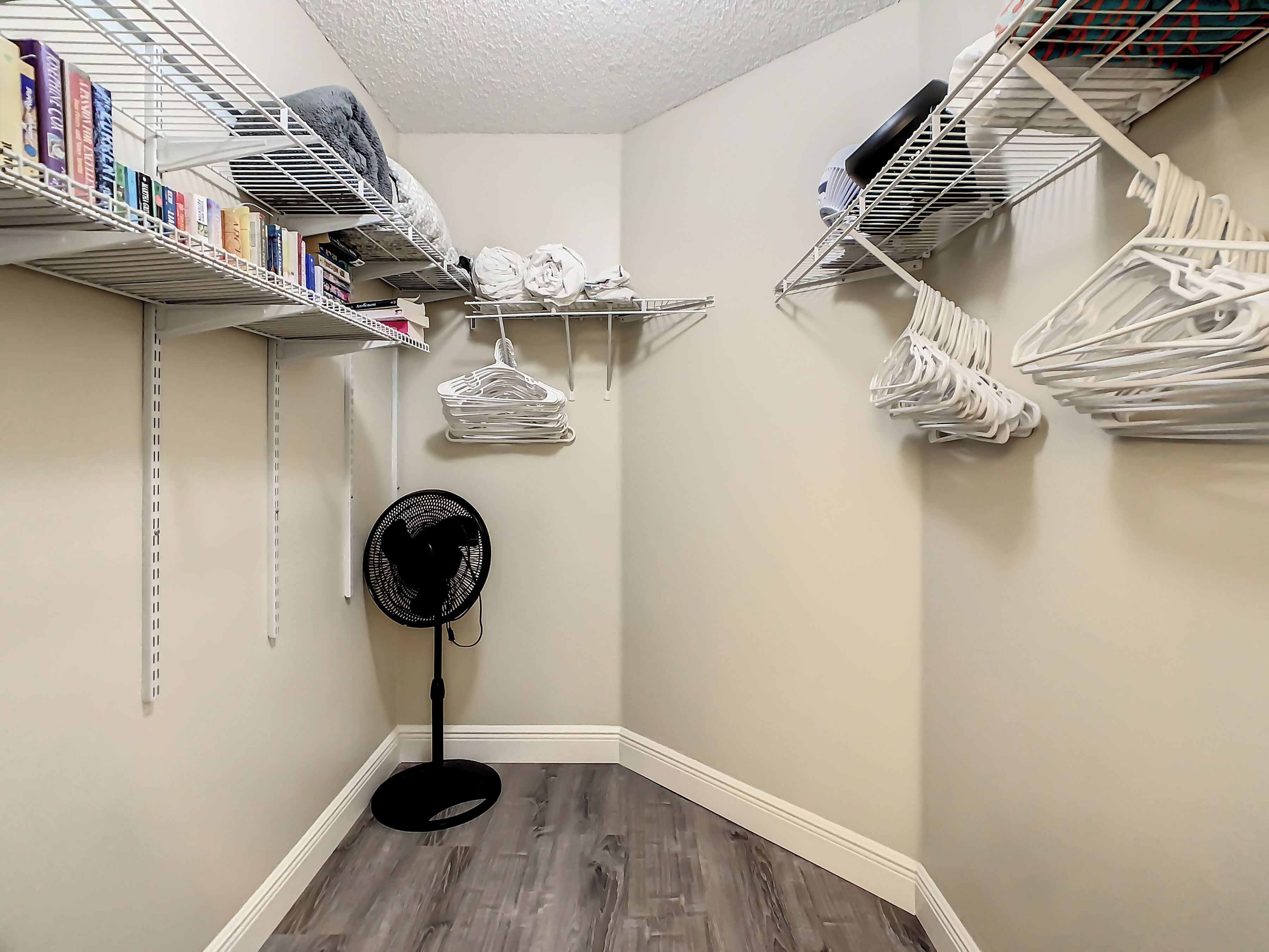 A corner closet with shelves for books and hanging clothes, a standing fan, and a wooden floor.