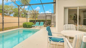 A covered outdoor pool area with lounge chairs and a table, surrounded by a fence and trees. - Southern Dunes Vacation Home (Haines City)