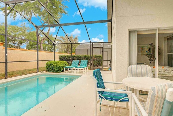 A covered outdoor pool area with lounge chairs and a table, surrounded by a fence and trees.