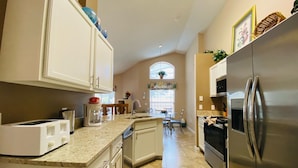 A kitchen with white cabinets, a stainless steel refrigerator, a coffee maker, and a dining area with chairs and a table. - Southern Dunes Vacation Home (Haines City)