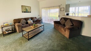 Sala de estar com sofá, mesa de centro e pequena mesa lateral. Há uma janela com cortinas, um quadro na parede e um aviso indicando disponibilidade de Wi-Fi.