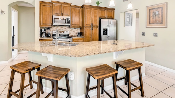 A kitchen with wooden cabinets, a stainless steel refrigerator, a microwave, and a granite countertop. There are three wooden stools at the island.