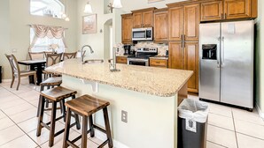 A kitchen with wooden cabinets, a stainless steel refrigerator, a granite countertop, and a dining area with chairs and a table.