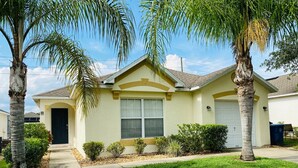 A single-story house with a light yellow exterior, a dark brown roof, and a front yard with a well-maintained lawn and palm trees. - Mike's Southern Dunes Vacation Home (Haines City)
