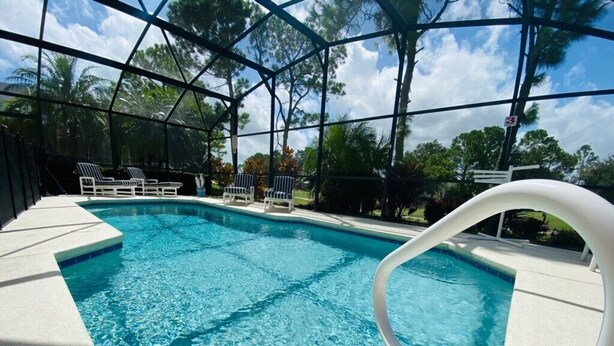 A clear blue pool with a white deck and lounge chairs, surrounded by a covered area with trees.
