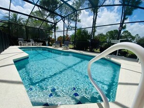 A clear blue pool with a white deck and lounge chairs, surrounded by a covered area with trees. - Villa Rosalie at Southern Dunes (Haines City)