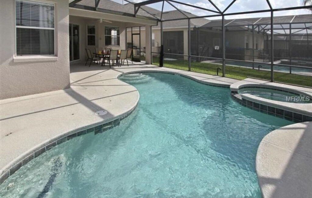 A swimming pool with a curved design, surrounded by a covered patio area with chairs and a table.