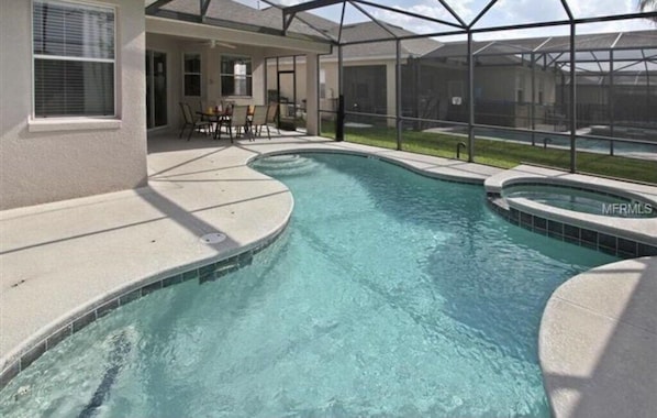 A swimming pool with a curved design, surrounded by a covered patio area with chairs and a table.