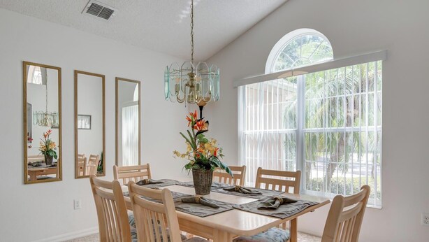 A dining area with a wooden table, chairs, and a chandelier. There are large windows with a view of greenery outside. - Maria's Southern Dunes Vacation Home (Haines City)
