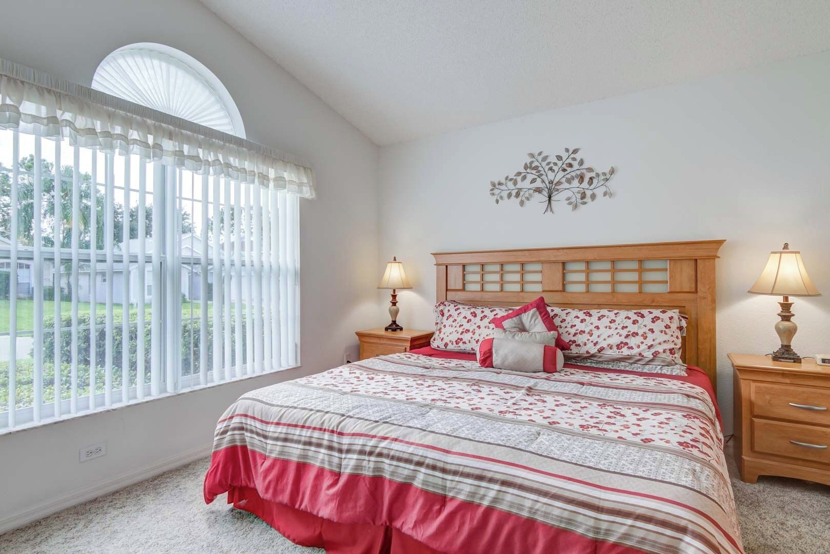 A bedroom with a large window, a wooden headboard, two bedside tables with lamps, and a decorative wall piece.