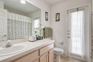 Une salle de bain avec un lavabo, un meuble-lavabo et des toilettes, le tout de couleur blanche. On trouve des objets décoratifs sur le meuble-lavabo, notamment un vase avec des fleurs et une petite boîte. La pièce est dotée d’un grand miroir, d’une fenêtre avec un rideau et d’une porte avec une poignée décorative.