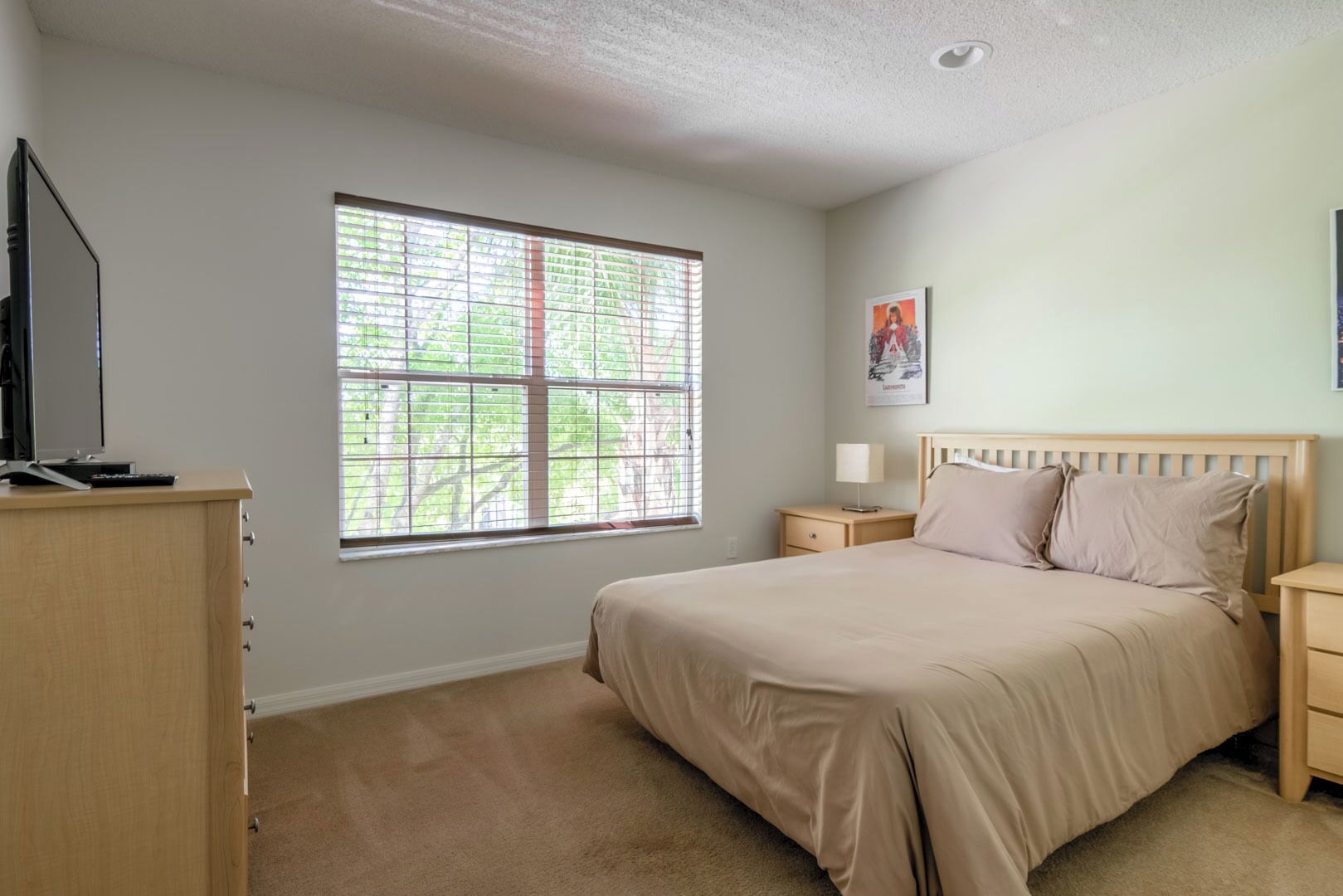 A bedroom with a bed, a nightstand, a dresser, a window, and a wall-mounted TV.