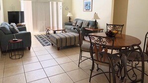 Ein geräumiges Wohnzimmer mit einer Couch, einem Esstisch mit Stühlen, einem Fernseher und einem Fenster mit Jalousien.