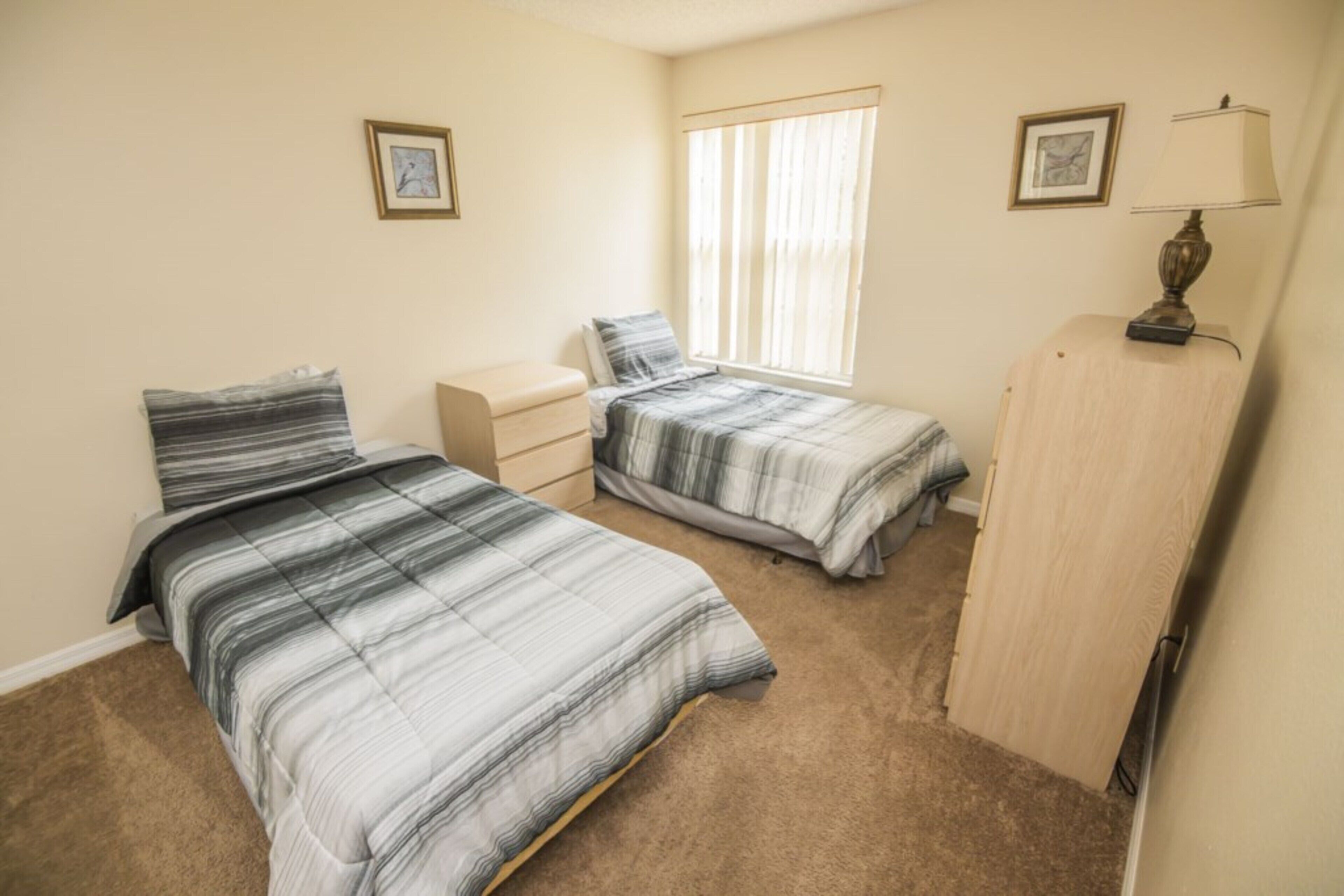 A bedroom with two beds, a dresser, a window with blinds, and two framed pictures on the wall.