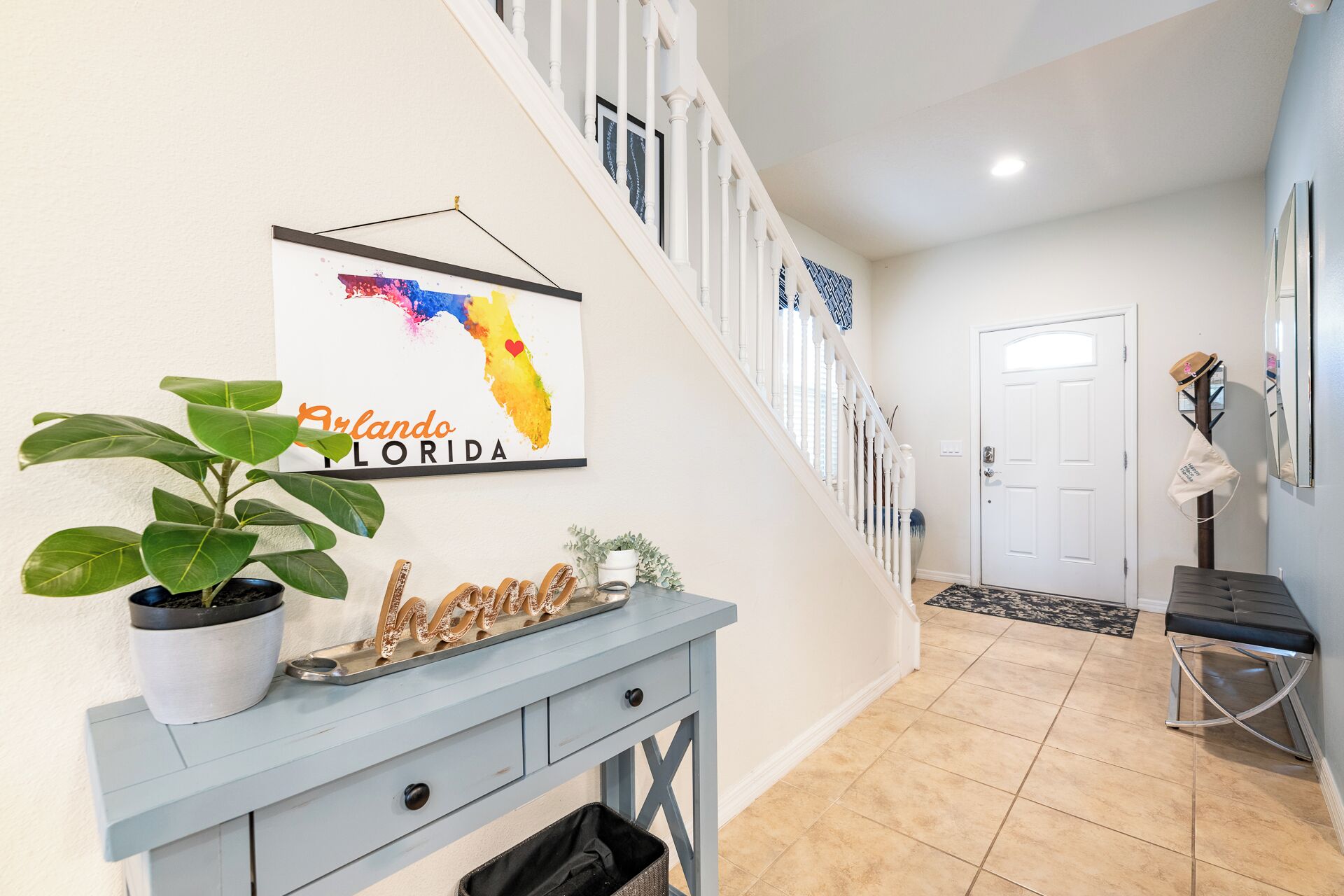 A hallway with a staircase, a framed sign reading "Orlando Florida", a potted plant, a decorative word "home", and a bench.