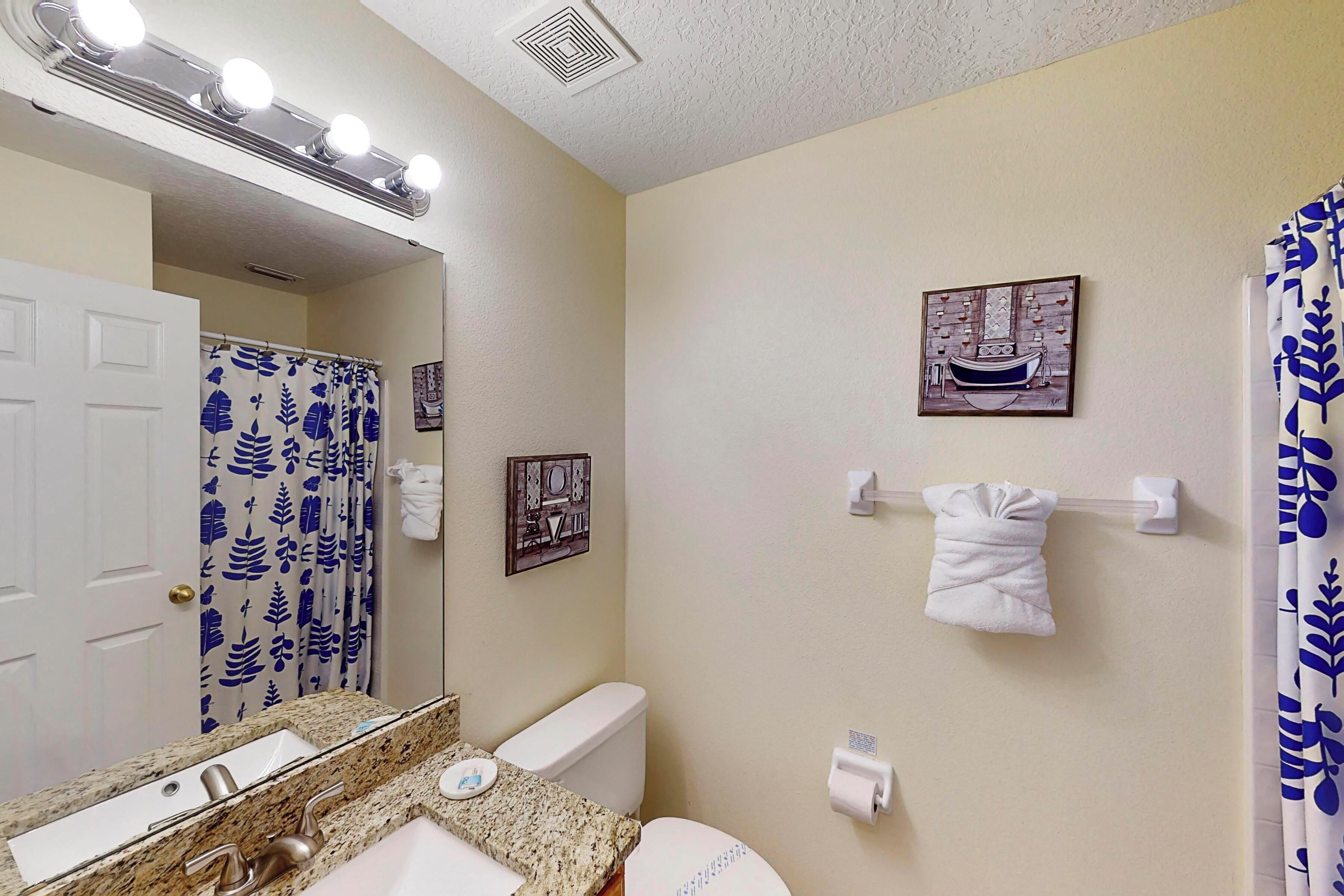 Une salle de bain avec un lavabo, un miroir et des toilettes. Il y a des lampes au-dessus du miroir, un porte-serviettes et un tableau au mur.