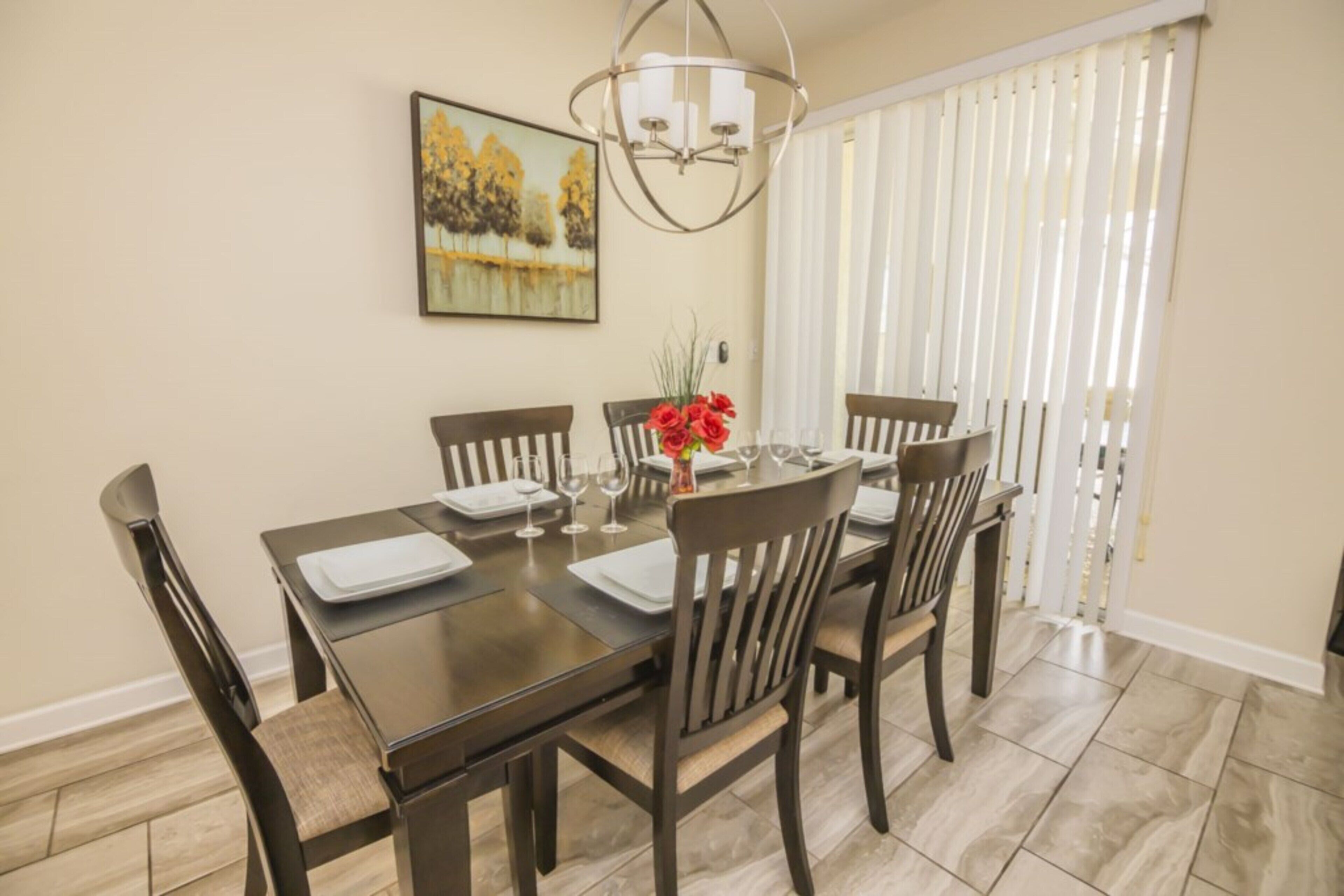 A dining area with a rectangular table set for four, surrounded by wooden chairs. A chandelier hangs above the table, and a painting is on the wall.