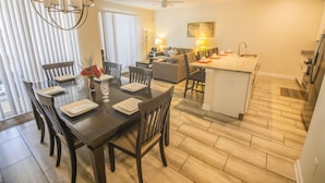 A modern kitchen with a central island, dining area with a dark wooden table and chairs, and a living area with a sofa and lamp. - Brenda's Storey Lake Resort Townhome (Kissimmee)