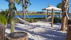 A beach scene with a hammock, a wooden fence, a gazebo, and a view of a body of water and buildings in the background. - Brenda's Storey Lake Resort Townhome (Kissimmee)