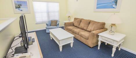 A room with a desk, a brown sofa, two white side tables, and a window with blinds.
