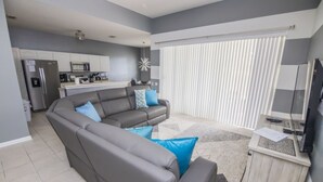 A modern living room with a grey sectional sofa, a flat-screen TV, and a kitchen area in the background. - Bernard's Windsor Hills Resort Vacation Home (Kissimmee)