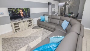 A modern living room with a large flat-screen TV, a grey sectional sofa, and a dining area in the background. - Bernard's Windsor Hills Resort Vacation Home (Kissimmee)