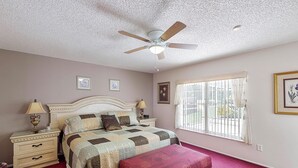 A bedroom with a bed, nightstands, a red ottoman, a ceiling fan, and a window with curtains.