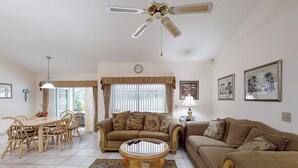 A living room with a dining table, two sofas, a coffee table, a ceiling fan, a clock, and framed pictures on the wall.