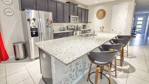 A kitchen with dark cabinets, a granite countertop, a refrigerator with a Mickey Mouse decal, a microwave, a sink, and bar stools. - Jodie's Luxury Solara Home (Kissimmee)