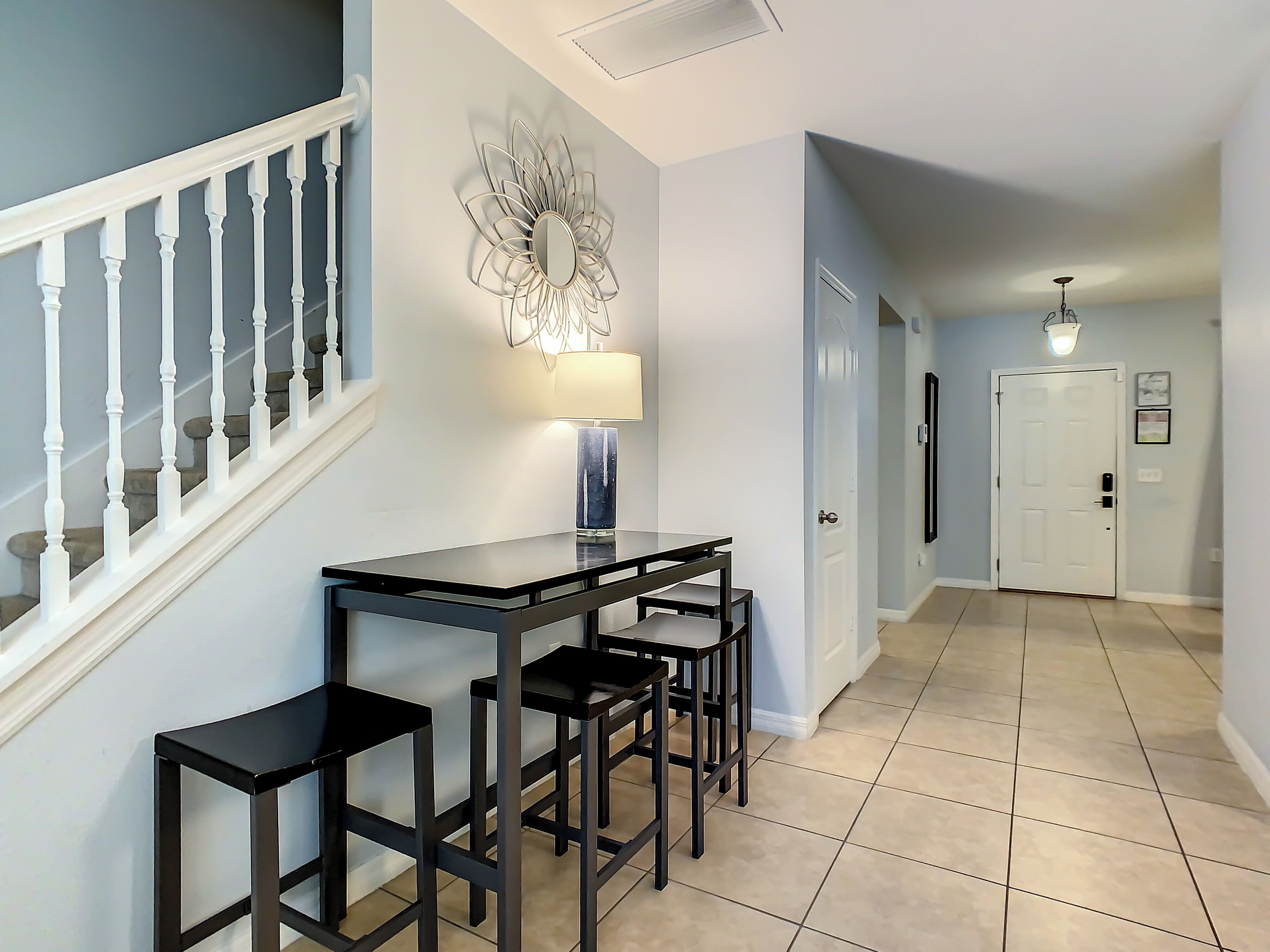 A hallway with a staircase, a dining table with chairs, a wall-mounted lamp, and a door at the end.