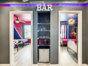 Ein Zimmer mit einem 'BAR'-Schild, einem Bett mit rosa Bettwäsche, einem Deckenventilator und einem an der Wand befestigten Regal mit verschiedenen Gegenständen.