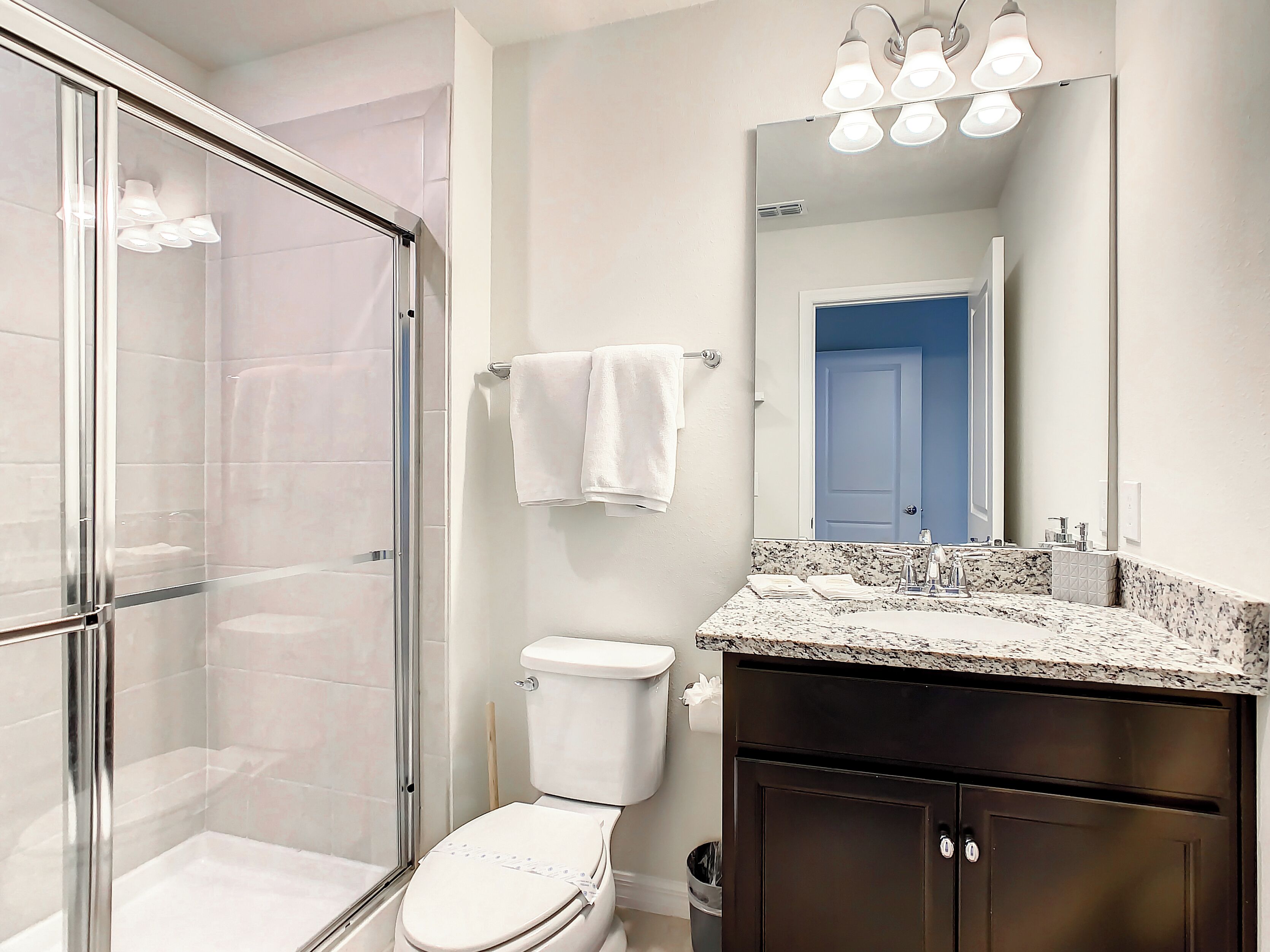 Une salle de bain moderne avec une douche à parois de verre, des toilettes blanches et un meuble-lavabo en bois foncé doté d’un plan de travail en granit.