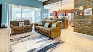 A living room with two sofas, a coffee table, and a TV. There is a kitchen in the background with wooden cabinets and a stone wall. - Comrow Castle Magical Getaway (Kissimmee)