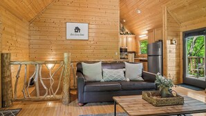 L’intérieur chaleureux d’un chalet, avec un revêtement mural en bois, un canapé en cuir noir, une table basse et un coin cuisine visible à travers une porte ouverte.